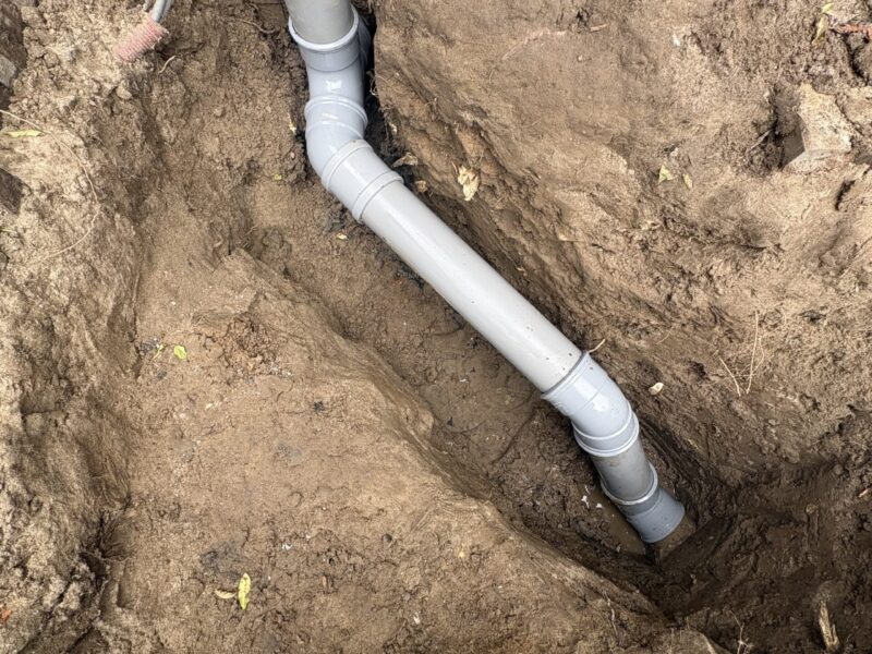 Nieuwe grijze PVC-rioolleiding met bochtstuk en inspectie-opening gelegd in uitgraving langs gevel in Barendrecht